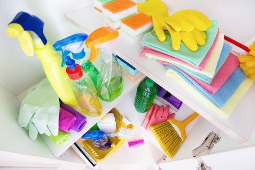 A collection of cleaning supplies and cleaning cloths arranged on a white surface, including spray bottles with yellow, blue, green, and orange nozzles, some containing cleaning liquids, alongside a pair of yellow rubber gloves, neatly folded colourful microfiber cloths in pastel shades, a small yellow dustpan with a brush, a purple sponge, and a metal clip, all positioned on a white shelf or tabletop. The background reveals a clean, bright environment, suggesting an indoor setting used for domestic or commercial cleaning tasks, with the items organized together. The scene emphasizes tools and materials commonly associated with cleaning or waste management contexts, and the overall composition reflects attention to orderliness and preparedness for cleaning activities, aligning with rubbish removal services offered by businesses like Business Waste Removal Southwark, serving the Southwark area in London postcode.