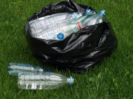 A black plastic rubbish bag placed on a grassy area, partially open at the top revealing clear plastic bottles with blue caps and one with a red ring, all of which appear to be discarded beverage containers. Two additional empty bottles are lying on the ground close to the bag, one near the front left and the other just beside the bag on the grass. The bottles are transparent with textured ridges along their bodies and are made of lightweight plastic. The scene is outdoors, with vibrant green grass in the background and foreground, suggesting a residential or public space in Southwark where rubbish removal services might be needed for garden or street cleanup. The lighting is natural, indicating daylight, and the overall setup emphasizes the typical waste collected and processed by companies like Business Waste Removal Southwark, focused on waste disposal and recycling within the local area. The image highlights the need for proper rubbish collection, especially of recyclable bottles, as part of effective waste management for Southwark businesses.